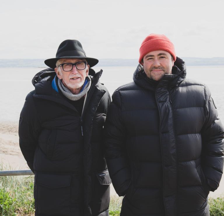 Two peope wearing black bomber coats and hats (one orange beanie and one black wide brimmed) standing with their backs to the sea