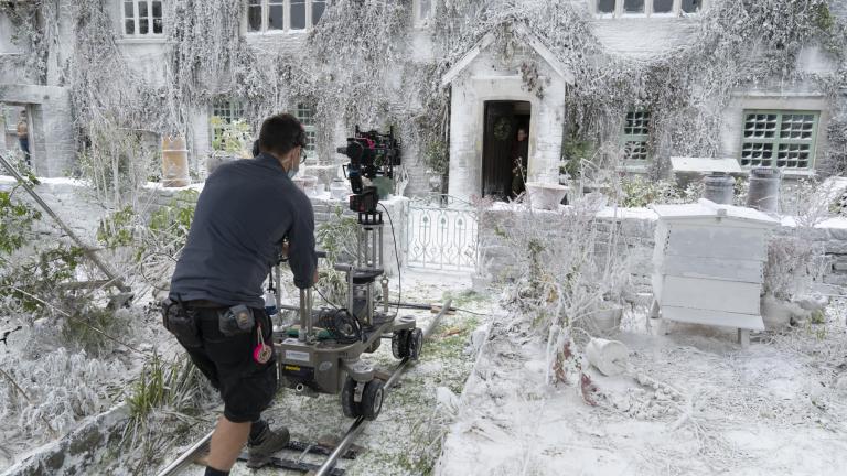 A person stands on a mini track holding a film camera. They are focusing on a white house covered in snow.