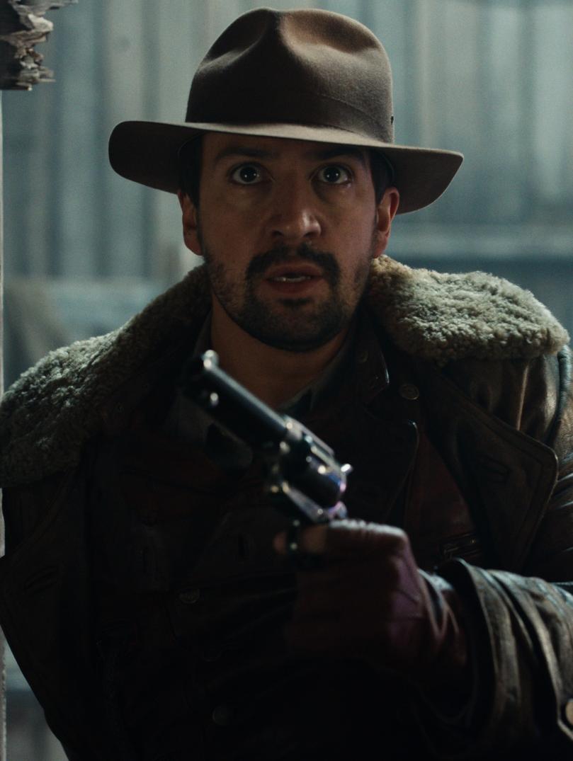 Close up still of Lee Scoresby (Lin-Manuel Miranda) from His Dark Materials (Series 2) (Bad Wolf). Lin-Manuel is wearing wearing a brown hat, brown jacket and is holding a metal handgun.