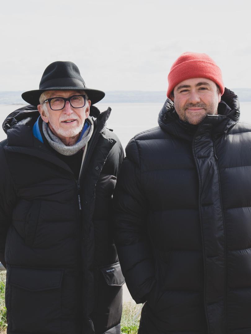 Two peope wearing black bomber coats and hats (one orange beanie and one black wide brimmed) standing with their backs to the sea