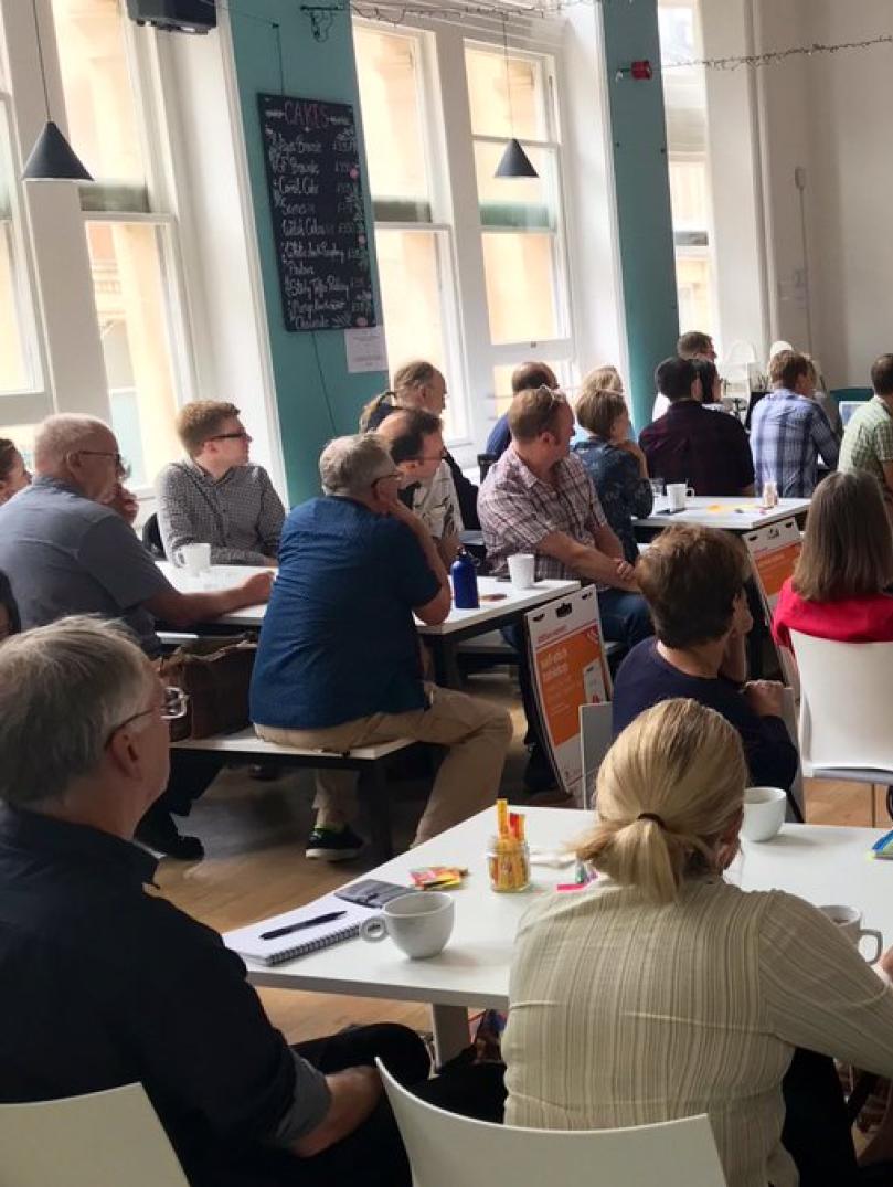 A large group of people sitting at white tables facing a projector screen as someone presents. The screen shows a background of colourful balloons with the word "Clwstwr" in white text in the middle.