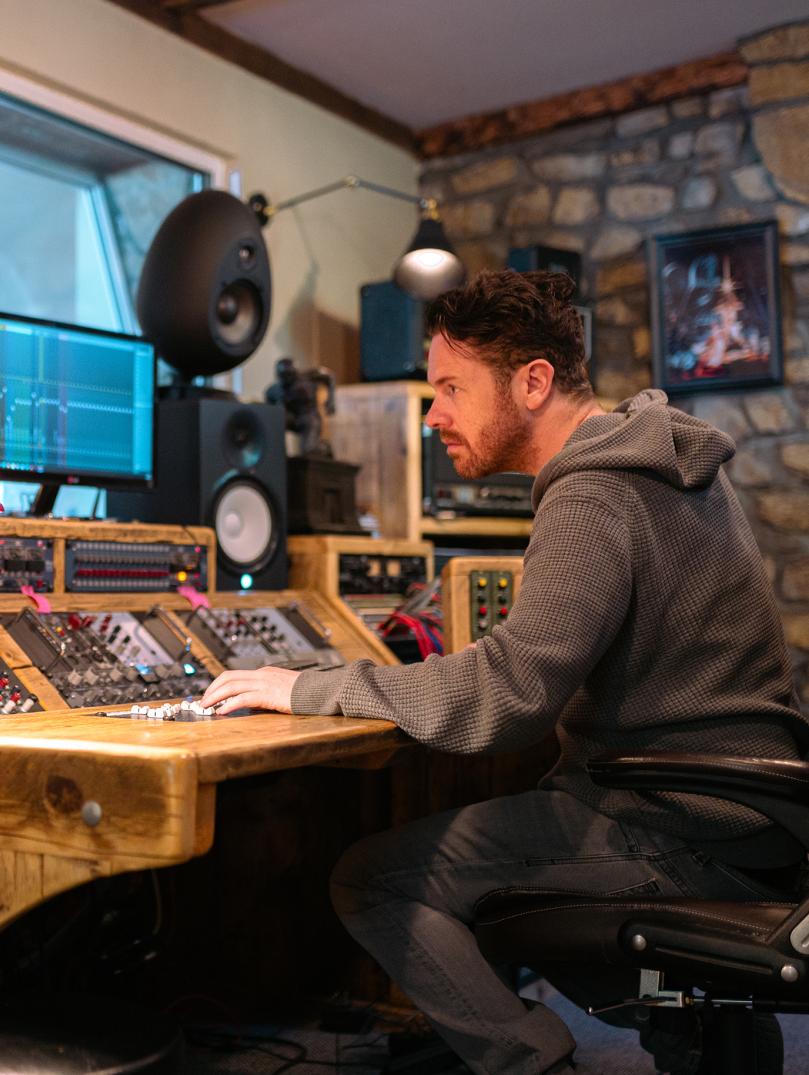 Jayce Lewis of Northstone Studios sits at a wooden mixing desk with monitors and large speakers in a dimly-lit studio.