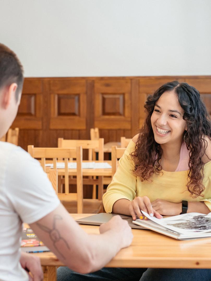 Close up shot of Yassmine from Bomper Studio talking to colleague. Yassmine is smiling, sitting at a wooden table wearing a yellow of the shoulder top.