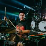 Medium shot of Ryan Richards playing drumps on a dimly lit stage. Ryan is wearing a black baseball cap and a black tshirt with an orange design.