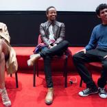 Three people sit together on red chairs. They are all looking to towards the right of the shot and laughing. The floor is red carpet and the wall behind is half black and half white.