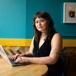 Yasmine has dark brown hair and sits at a table on a laptop and smiles at the camera
